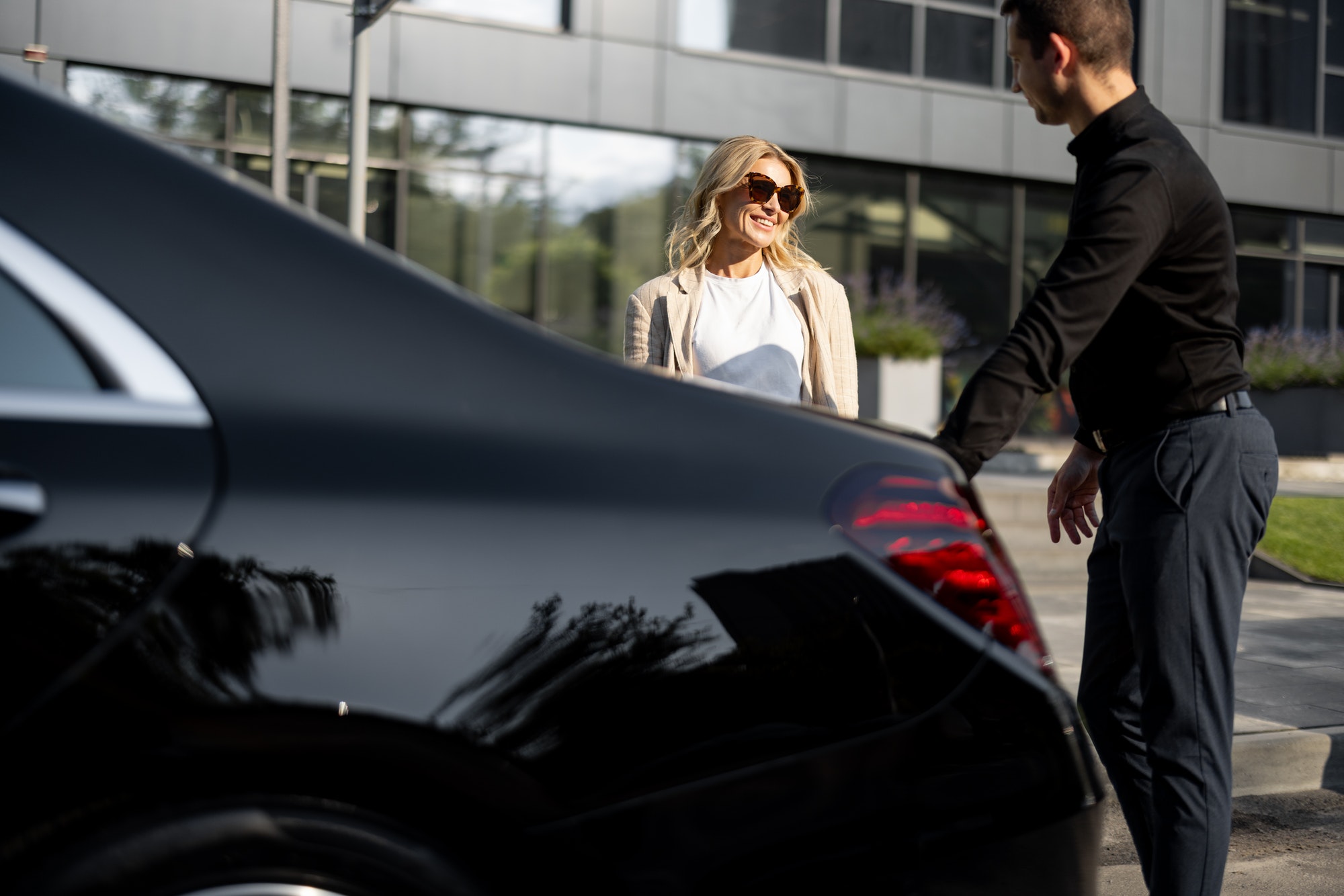 Une femme d'affaires attend pendant que le chauffeur prépare une valise dans le coffre de la voiture, en utilisant un service de taxi de luxe lors d'un voyage d'affaires.