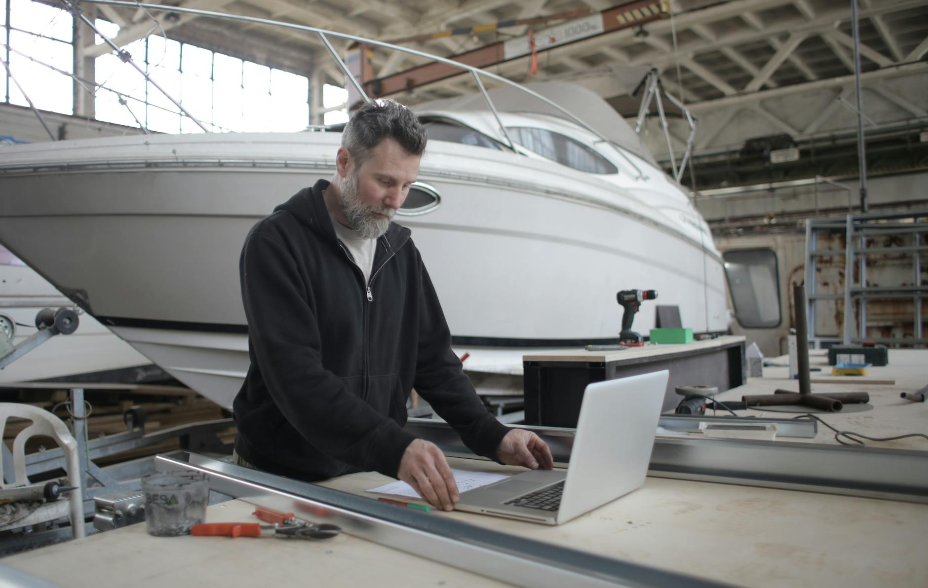 Travailleur adulte réfléchi utilisant un ordinateur portable tout en travaillant avec des pièces métalliques près du bateau en atelier