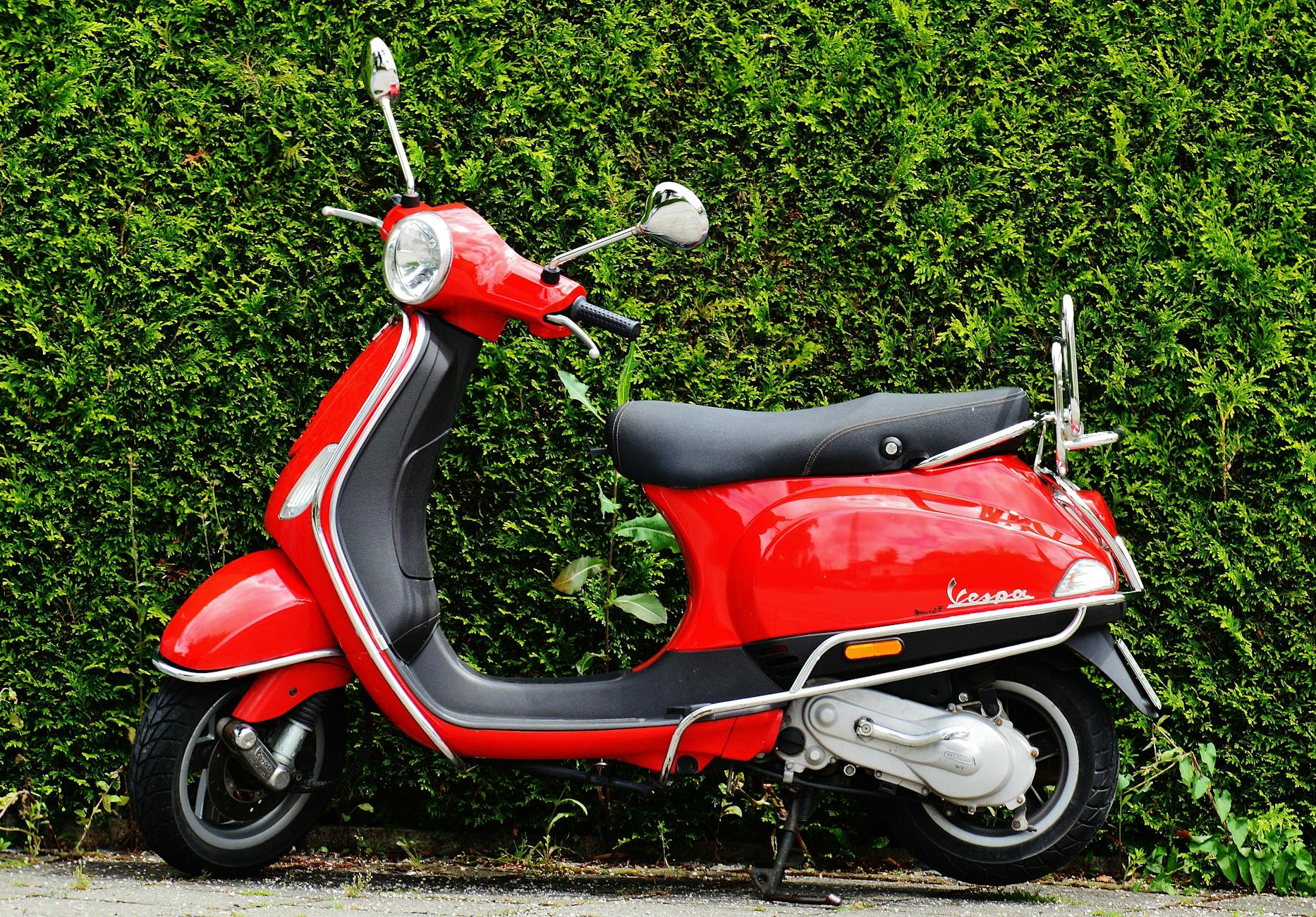 Scooter Vespa rouge vif garé contre une haie verte luxuriante, présentant un contraste dynamique.