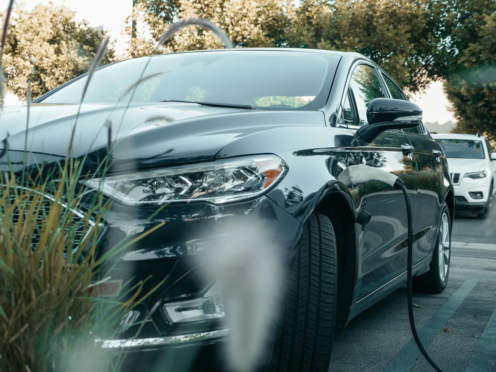 Une voiture hybride noire charge dans une station extérieure entourée de verdure.