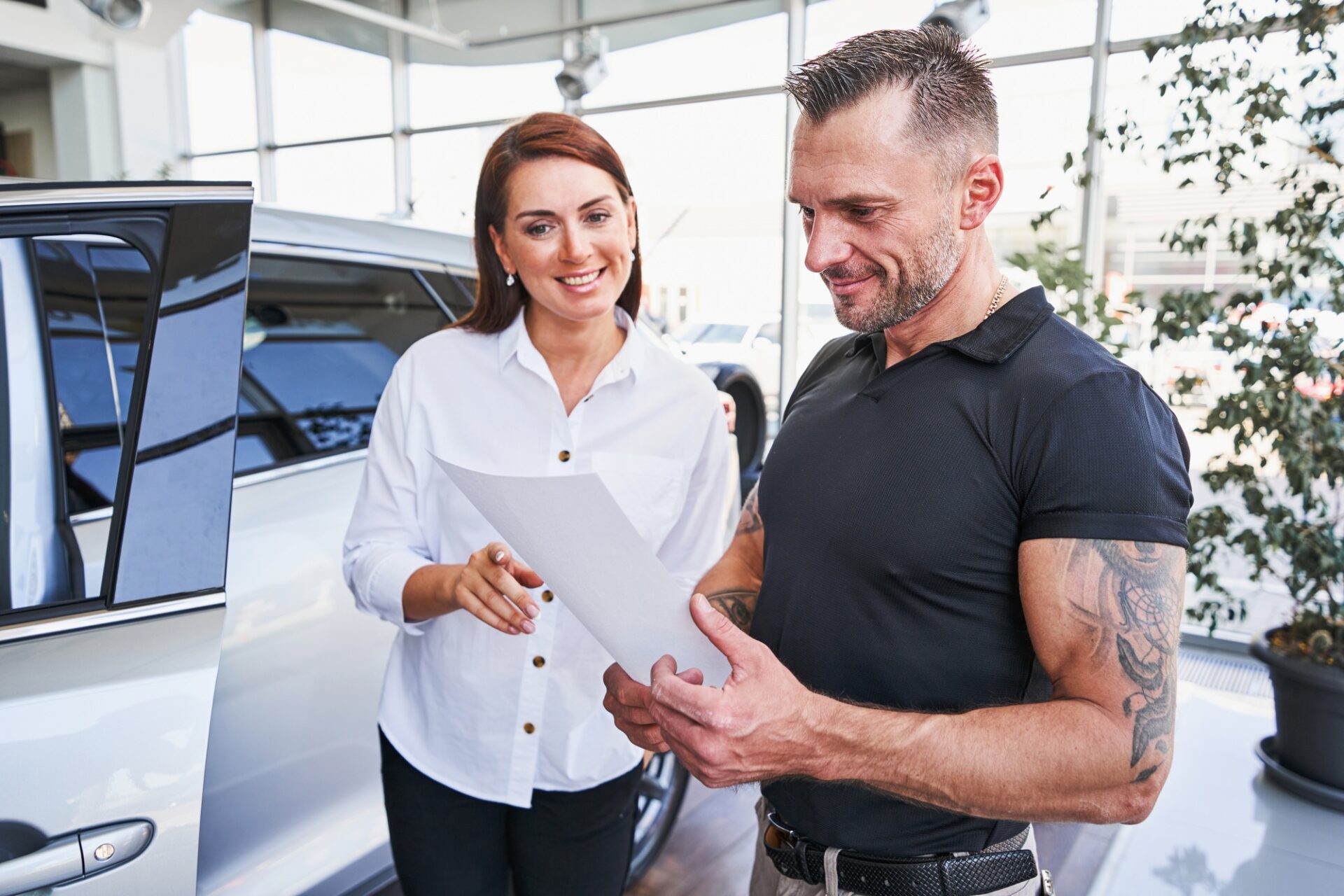 Une vendeuse amicale de joie pointant vers une feuille de papier entre les mains d'un homme en salon de voiture