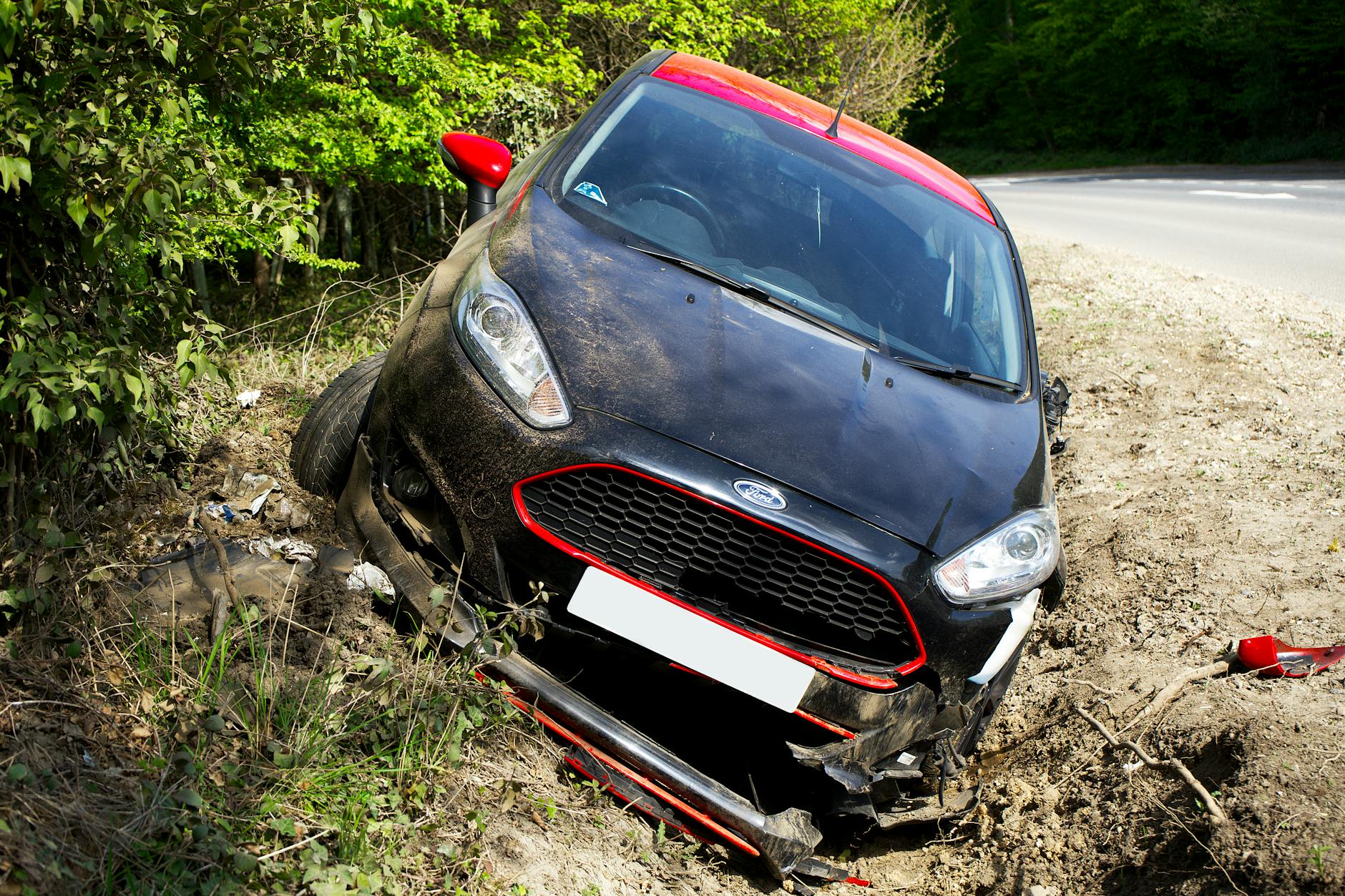 Une voiture noire, apparemment une Ford, s'est écrasée et a été endommagée dans un fossé en bord de route entouré de verdure.