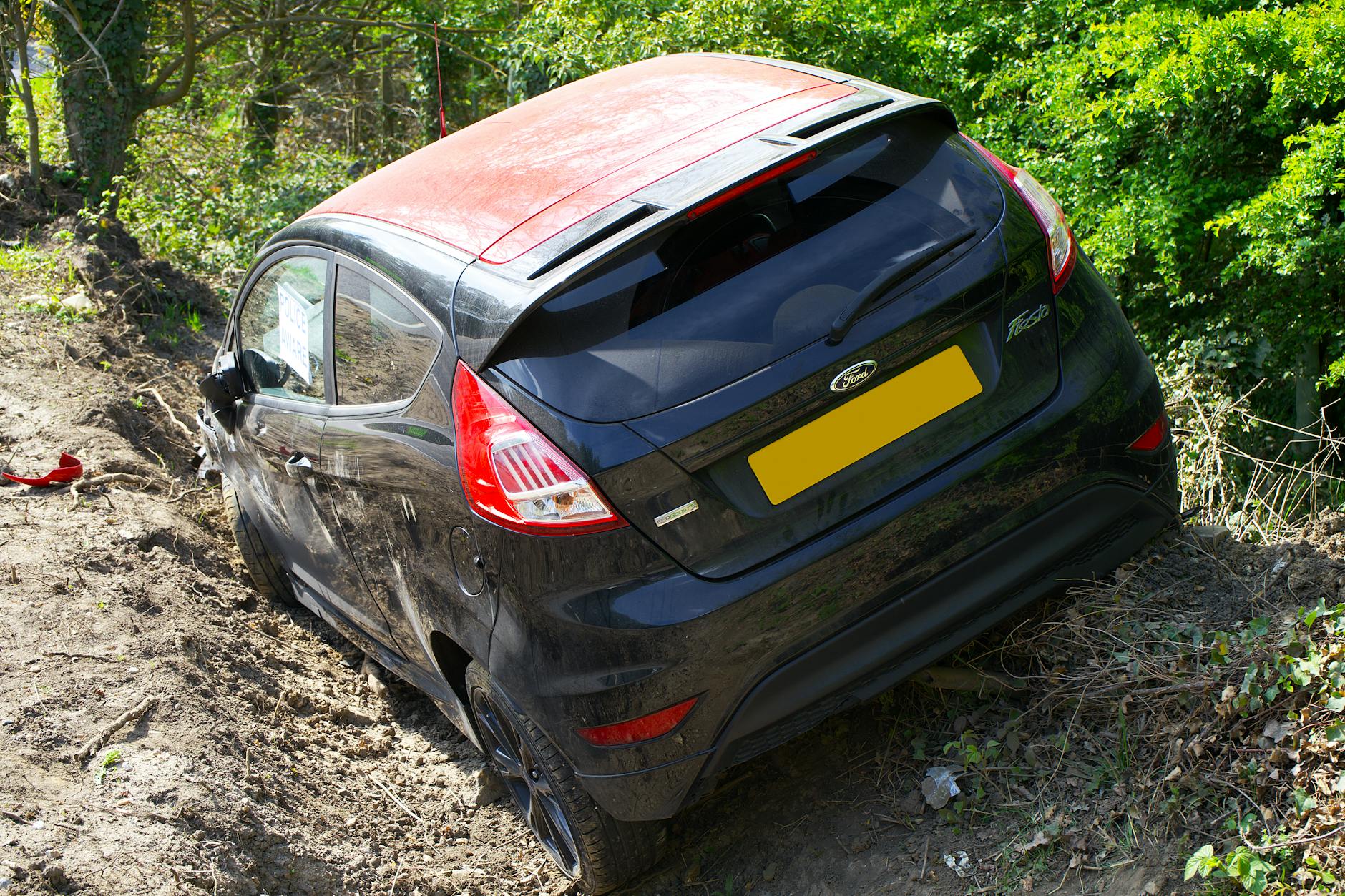 Ford Fiesta endommagée au bord d'un chemin de terre, entourée de feuillage vert, indiquant une scène d'accident.